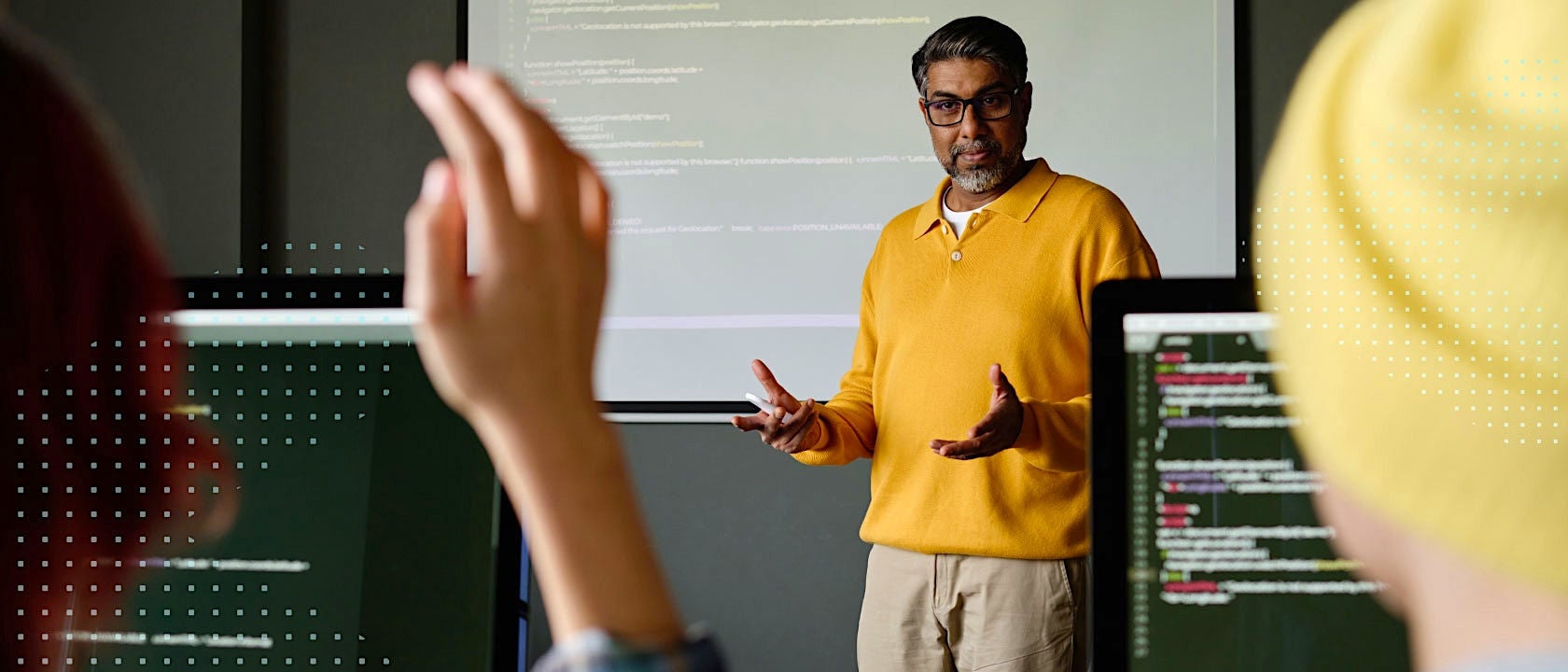 Photo of a man in a yellow shirt in front of a white board screen, with two students on computers in the foreground